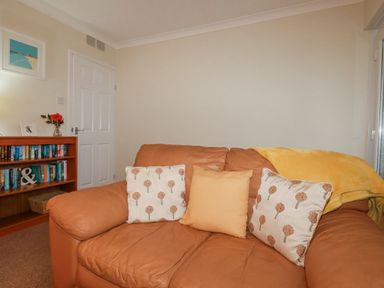 A living room with a brown leather sofa with cushions a yellow blanket a bookshelf with books and a vase with flowers at Garden View in Quintrell Downs near Newquay