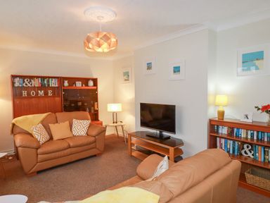 A living room with two brown sofas a wooden TV stand with a television a wooden bookcase with books and lamps at Garden View in Quintrell Downs near Newquay