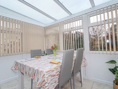 A dining area with a table covered by a patterned tablecloth and four chairs in a conservatory with vertical blinds at Garden View in Quintrell Downs near Newquay