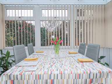 A dining table with six grey chairs and tulips in a vase in a room with large windows and vertical blinds at Garden View in Quintrell Downs near Newquay