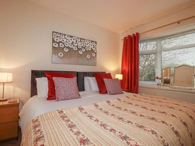 A bedroom with a bed featuring red and floral pillows a floral quilt bedside tables with lamps red curtains and a window at Garden View in Quintrell Downs near Newquay