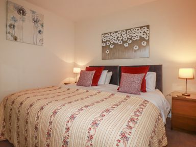 A bedroom with a double bed red and patterned pillows two bedside tables with lamps and floral paintings on walls at Garden View in Quintrell Downs near Newquay