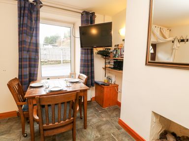 A dining area with a wooden table set for three near a window with blue checked curtains and a wall-mounted TV at Hunter Cottage in Haltwhistle near Hadrians Wall