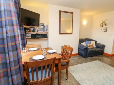 A dining table with four plates and glasses near a wall-mounted TV and shelves with books and a stereo and a small sofa with cushions in a living area at Hunter Cottage in Haltwhistle near Hadrians Wall