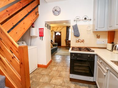 A kitchen with a gas stove oven under wooden stairs and a view into a dining area and entrance at Hunter Cottage in Haltwhistle near Hadrians Wall