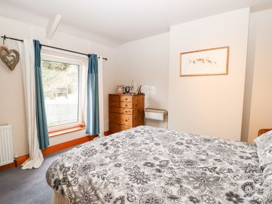 A bedroom with a bed covered in floral bedding a wooden chest of drawers a window with blue and white curtains and a framed picture on the wall at Hunter Cottage in Haltwhistle near Hadrians Wall