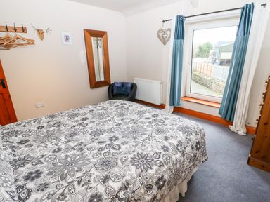 A bedroom with a bed covered in floral patterned bedding a large window with curtains a black chair a wall mirror and wooden furniture at Hunter Cottage in Haltwhistle near Hadrians Wall
