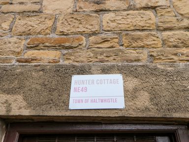 A white sign with text hunter cottage ne49 town of haltwhistle mounted on a stone wall at Hunter Cottage in Haltwhistle near Hadrians Wall