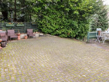 An outdoor patio with brick paving surrounded by chairs, a green fence, potted plants, and dense bushes at Hunter Cottage in Haltwhistle near Hadrians Wall