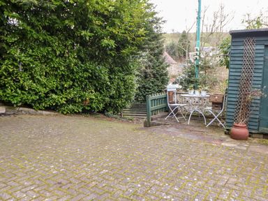 An outdoor patio area with a small round table and three chairs next to a green wooden shed and large leafy bushes at Hunter Cottage in Haltwhistle near Hadrians Wall