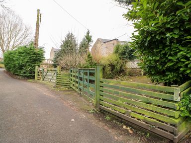 A country road beside a wooden green fence and gate with shrubs and stone buildings in the background at Hunter Cottage in Haltwhistle near Hadrians Wall