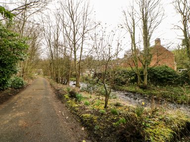 A narrow paved path alongside a small river with houses and trees nearby at Hunter Cottage in Haltwhistle near Hadrians Wall