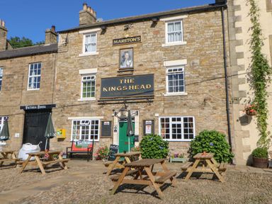 An outdoor seating area with wooden picnic tables in front of a stone building with signs reading The Kings Head and Victoria House at The Forge in Allendale