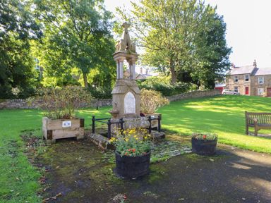 A stone monument with plaques in a grassy park with flower pots benches and houses in the background at The Forge in Allendale