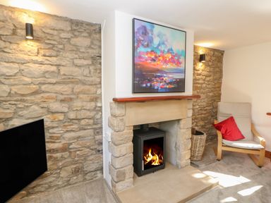 A room with a stone fireplace with a fire burning a painting above it a wooden chair with a red cushion and a basket at The Forge in Allendale