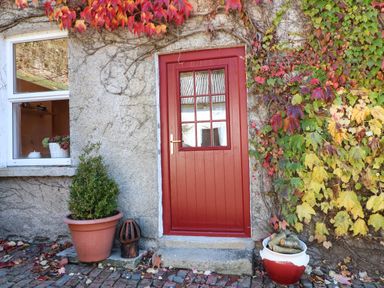 A red door with glass panels surrounded by a stone wall with autumn leaves and potted plants at Carley's Bridge House in Carley's Bridge near Enniscorthy County Wexford