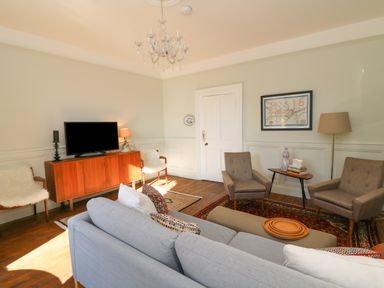 A living room with a gray sofa two armchairs a wooden cabinet with a television and a small table with books and a lamp at Carley's Bridge House in Carley's Bridge near Enniscorthy County Wexford