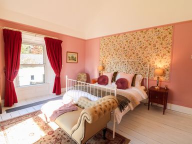 A bedroom with a white metal bed frame pink walls red curtains and a beige chaise lounge at Carley's Bridge House in Carley's Bridge near Enniscorthy County Wexford