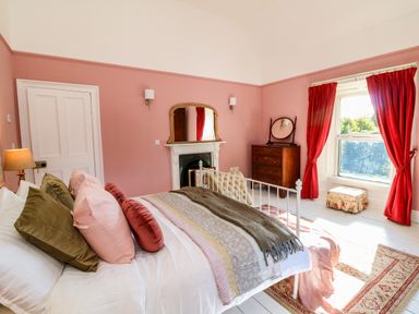 A bedroom with a bed pillows a fireplace a chest of drawers a window with red curtains and an ottoman at Carley's Bridge House Carley's Bridge near Enniscorthy County Wexford