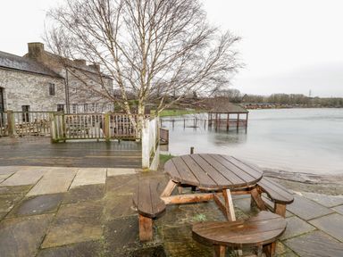 An outdoor seating area with wooden picnic tables and benches by a lake and stone buildings at Doormouse Lodge South Lakeland Leisure Village