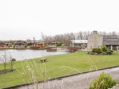 A lakeside area with a stone building a gazebo a bench and wooden cabins across the water at Doormouse Lodge South Lakeland Leisure Village