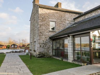 A stone building with large windows next to a paved walkway and grass near a body of water at Doormouse Lodge in South Lakeland Leisure Village