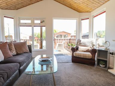 A living room with a sofa and armchair next to a glass coffee table and large windows at Doormouse Lodge in South Lakeland Leisure Village