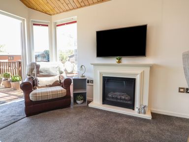 A living room with an armchair beside a fireplace and a mounted television at Doormouse Lodge South Lakeland Leisure Village