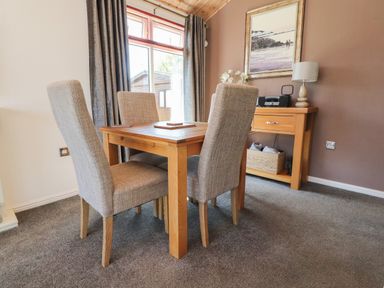A dining area with a wooden table and four fabric chairs next to a sideboard with a lamp radio and framed picture at Doormouse Lodge in South Lakeland Leisure Village