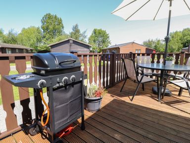 An outdoor deck with a barbecue grill and a round table with chairs and an umbrella at Doormouse Lodge South Lakeland Leisure Village