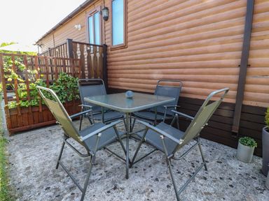Outdoor patio area with a square glass table and four metal chairs next to a wooden cabin at Doormouse Lodge in South Lakeland Leisure Village