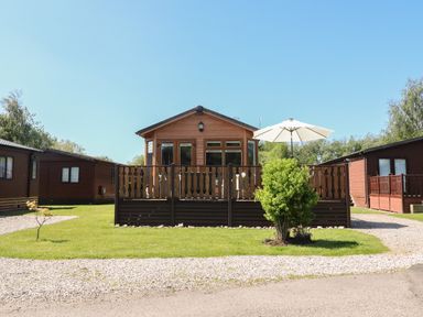 A small wooden lodge with a deck and white patio umbrella on a grassy plot at Doormouse Lodge in South Lakeland Leisure Village