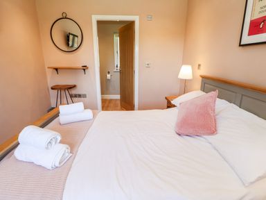 A bedroom with a bed with white sheets and pink pillow towels on bed a wooden side table with a lamp a wooden stool and a round mirror on the wall at Rising Sun in Kirkbymoorside