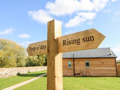 A wooden signpost with directions to Peep o day and Rising sun near a wooden building and stone wall in Kirkbymoorside