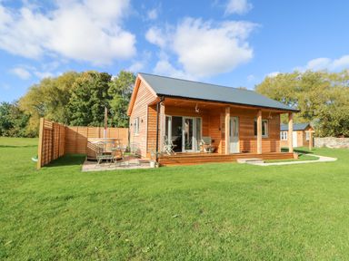 A wooden cabin with a front porch and patio furniture on a lawn with trees in the background at Rising Sun in Kirkbymoorside