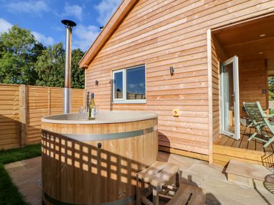 An outdoor hot tub with wine bottle and glasses next to a wooden cabin with open door and chairs at Rising Sun in Kirkbymoorside