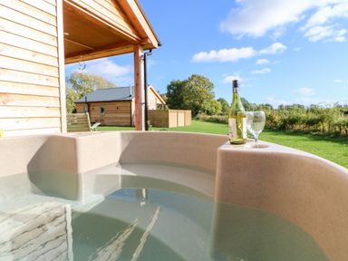 An outdoor hot tub with a bottle of wine and glass on the edge near wooden cabins and green grass at Rising Sun in Kirkbymoorside