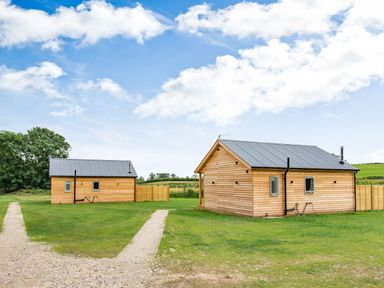 Two wooden cabins with metal roofs on a grassy field with a pathway and trees in the background at Peep o Day in Kirkbymoorside