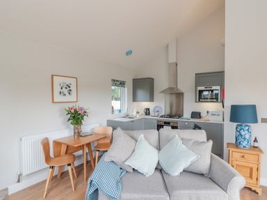 A compact kitchen and dining area with a small wooden table and chairs a grey sofa with cushions and a wooden side table with a blue lamp at Peep o Day in Kirkbymoorside