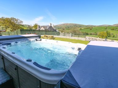 An outdoor hot tub with bubbling water and part of its cover open in a garden with a picnic table and green hills in the background at Bwthyn Y Bugail in Pedairffordd near Llanrhaeadr-Ym-Mochnant