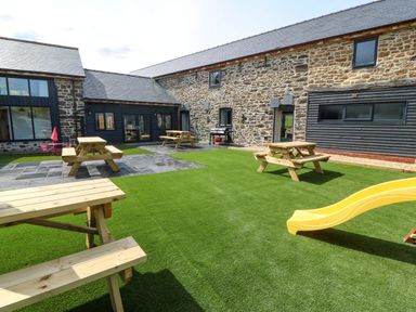 An outdoor courtyard with wooden picnic tables a yellow slide and stone buildings at Bwthyn Y Bugail Shepherd's Cottage in Pedairffordd near Llanrhaeadr-Ym-Mochnant