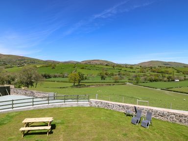 A grassy garden with a wooden picnic table and four lounge chairs overlooking green fields and hills at Bwthyn Y Bugail Shepherd's Cottage in Pedairffordd near Llanrhaeadr-Ym-Mochnant