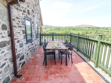 An outdoor patio with a table and six chairs next to a stone wall and green railing overlooking fields at Y Granar Llanrhaeadr-Ym-Mochnant