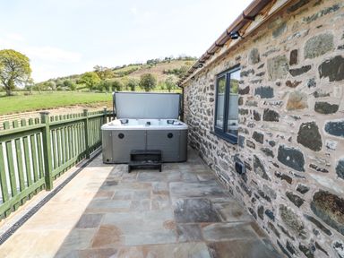 An outdoor patio area with a hot tub next to a stone wall and green railing with countryside view at Y Granar Llanrhaeadr-Ym-Mochnant