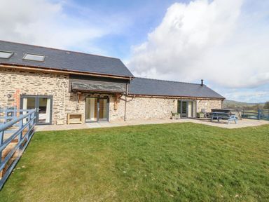 An outdoor area with a stone building with multiple doors and windows a grass lawn and a patio with a picnic table at Y Granar The Granary in Llanrhaeadr-Ym-Mochnant