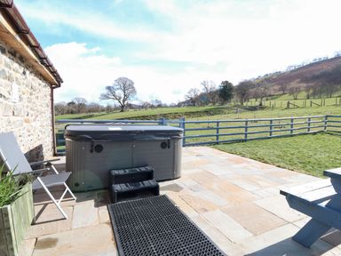 An outdoor patio with a hot tub chairs and a picnic table next to a stone building with fields and hills in the background at Y Granar Llanrhaeadr Ym Mochnant
