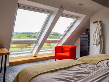 A bedroom with two beds a red armchair and large slanted windows at Y Granar The Granary in Llanrhaeadr-Ym-Mochnant