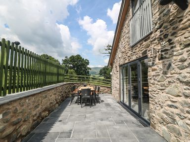 An outdoor patio area with a stone wall and wooden fence a table with six chairs and a bottle on the table at Y Stabl The Stable in Llanrhaeadr Ym Mochnant
