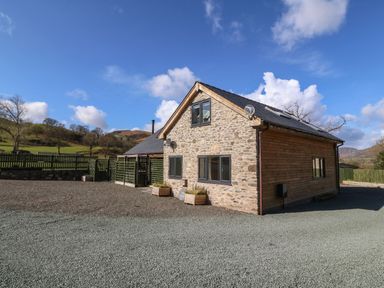 A stone and wood house with gravel driveway and green hills in the background at Y Stabl The Stable in Llanrhaeadr-Ym-Mochnant
