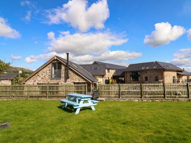A stone building with a wooden fence and a blue picnic table on green grass at Y Stabl The Stable in Llanrhaeadr-Ym-Mochnant